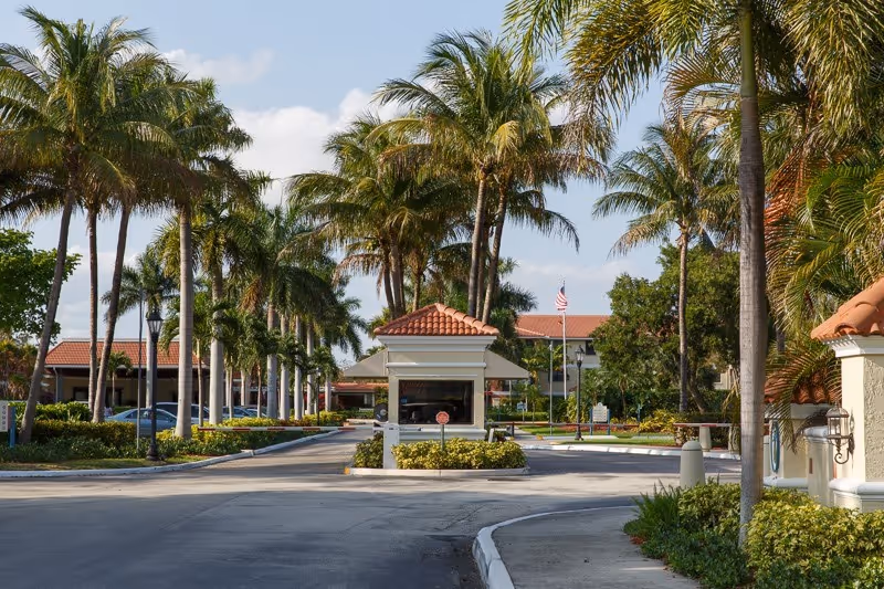 Entrance driveway to a senior living facility with a small guardhouse in the center, lined with tall palm trees and landscaped greenery on both sides. The building with a red-tiled roof is visible in the background under a partly cloudy sky.