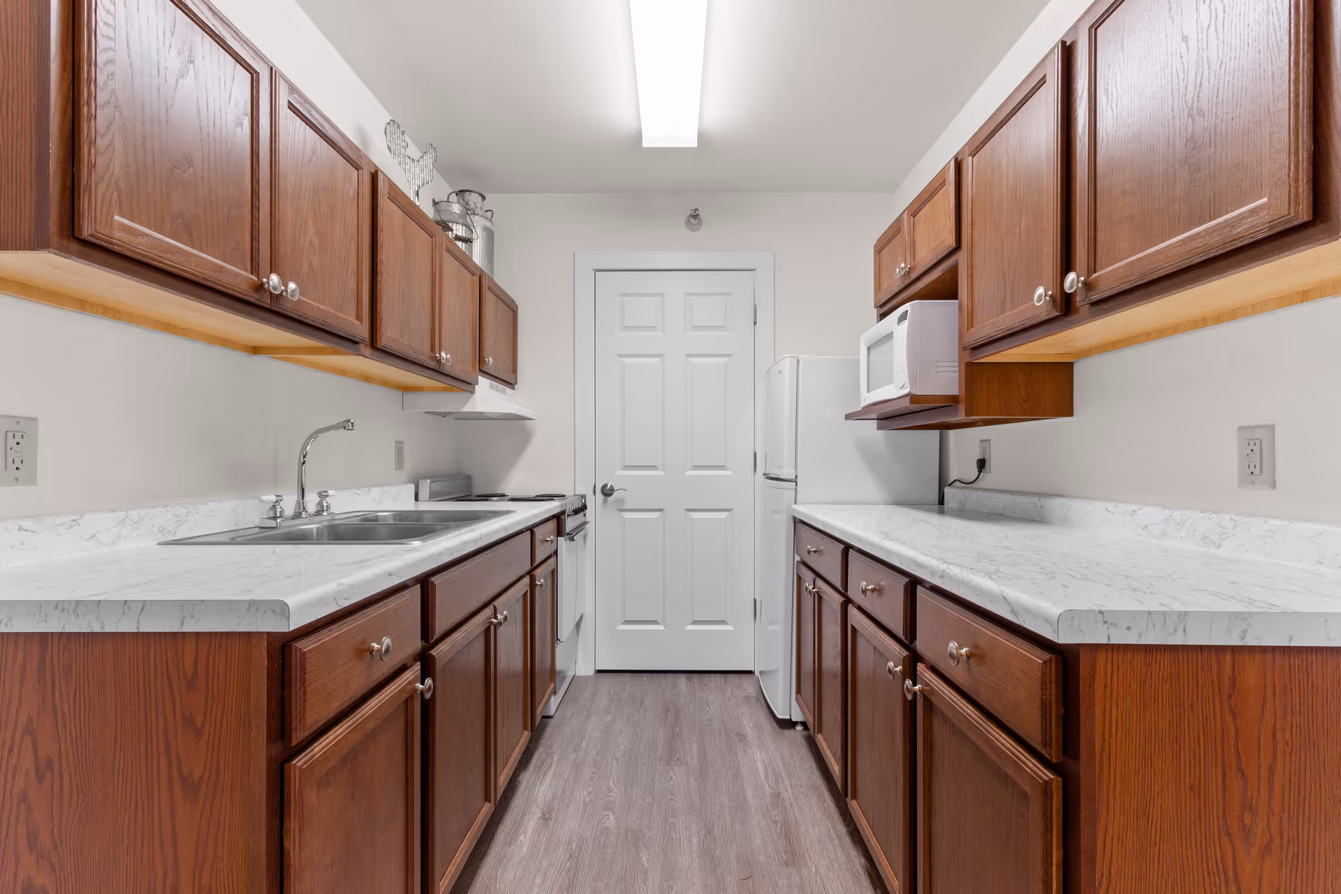 Narrow galley kitchen with dark wood cabinets, white countertops, a double sink on the left, and a refrigerator and microwave at the far end.