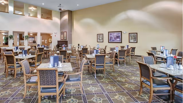 A spacious dining room with multiple square tables and wooden chairs arranged neatly. Each table has place settings including cups, napkins, and condiments. The room has a patterned carpet, light-colored walls with framed artwork, and a high ceiling with recessed lighting. There are open windows to an adjacent room on the left side.