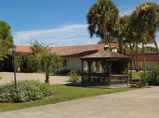 Front courtyard with a wooden gazebo, palm trees, and a low stucco building under a blue sky.