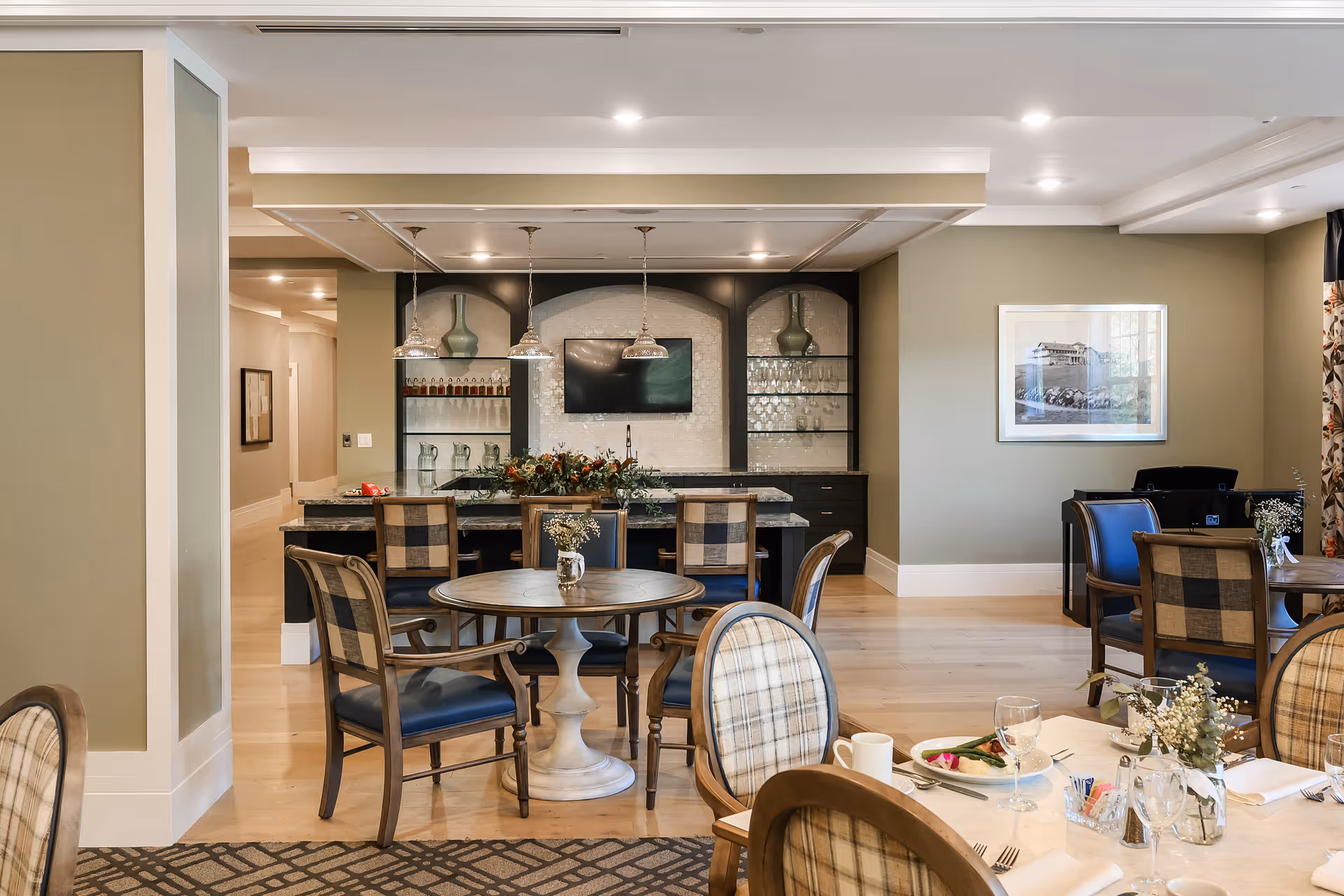 A cozy dining area in a senior living facility featuring round tables with floral centerpieces and cushioned chairs. In the background, there is a bar area with pendant lights, glass shelves holding glassware and decorative vases, and a mounted flat-screen TV. The room has light-colored walls, wooden flooring, and framed artwork on the wall.