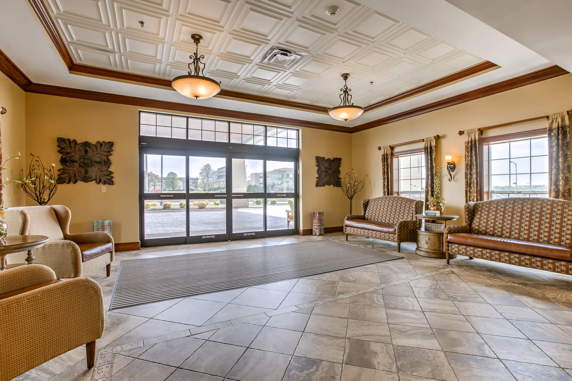 A spacious and well-lit lobby area with large glass entrance doors, beige tiled floor, and comfortable seating including armchairs and patterned sofas. The room features decorative wall art, large windows with curtains, and elegant ceiling lights with a coffered ceiling design.