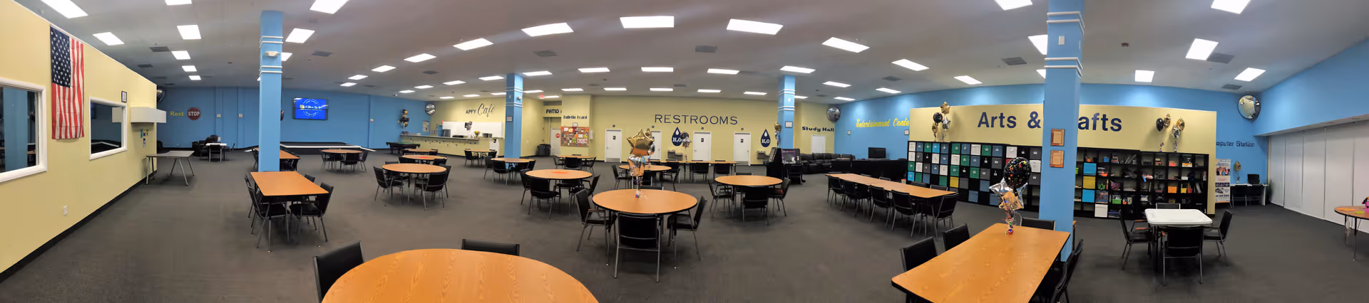 Panoramic view of a large, well-lit multipurpose room in an adult daycare and rehab center. The room has multiple round and rectangular tables with chairs arranged around them. The walls are painted in light yellow and blue, with signs indicating restrooms and areas for arts and crafts. There are shelves filled with colorful bins and supplies on one side, and an American flag is displayed on the left wall. The ceiling has recessed lighting, and the floor is carpeted.