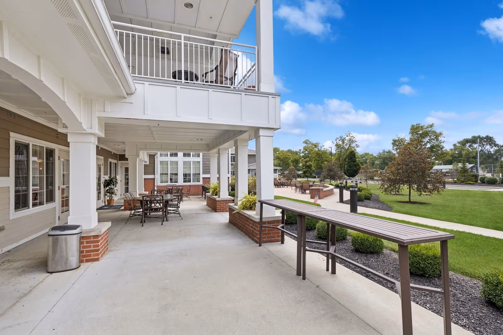 Outdoor covered patio area with tables and chairs, adjacent to a building with large windows. There is a balcony above with additional seating. The patio overlooks a landscaped garden with green grass, trees, and a clear blue sky.