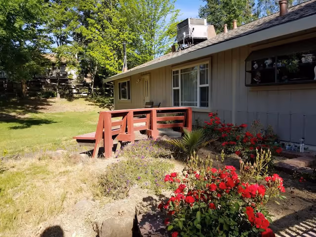 Exterior view of a single-story building with beige siding and a red wooden ramp leading to the entrance. The foreground features a garden with red flowers and other plants, and there are trees and grass surrounding the building under a clear blue sky.