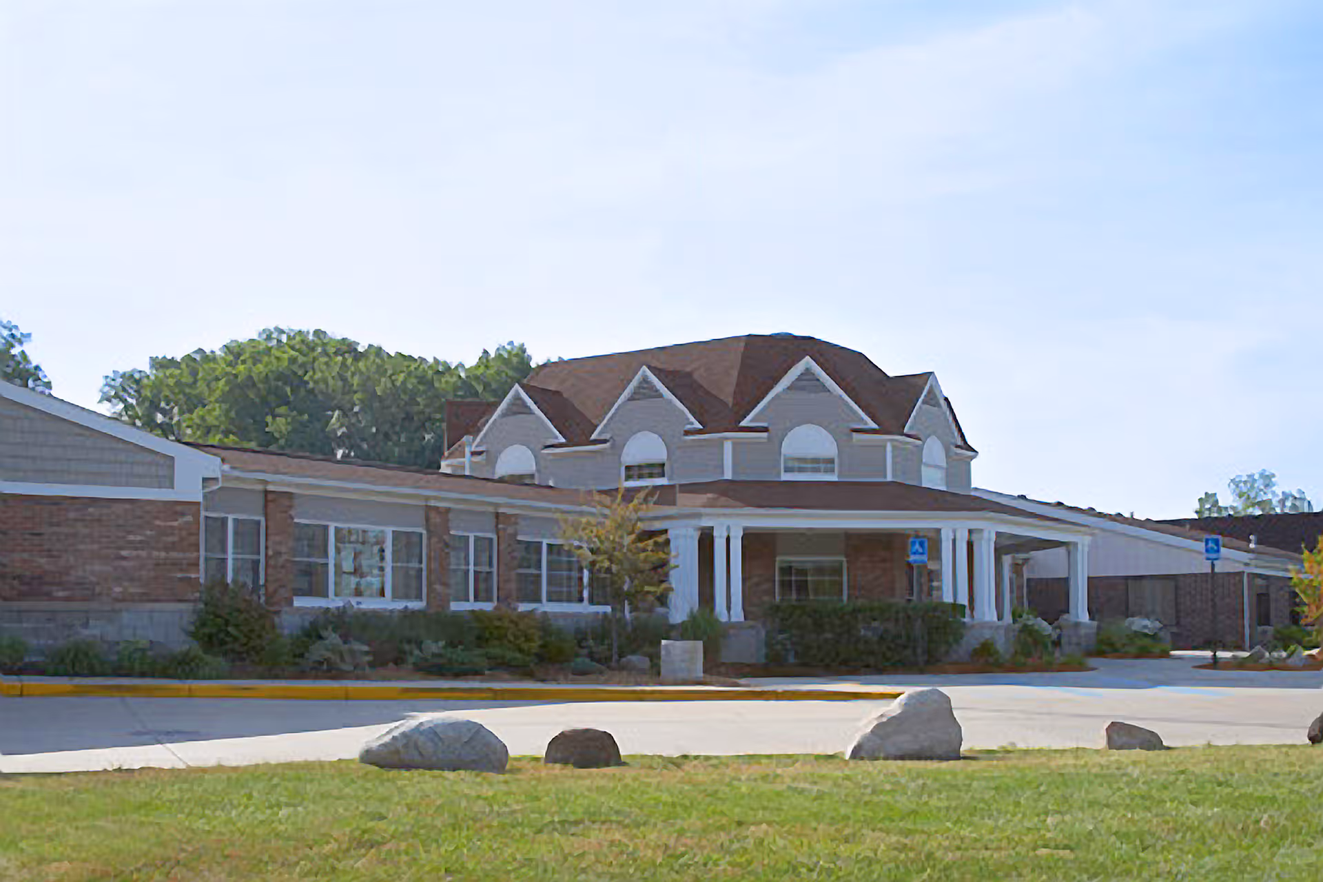 Exterior view of a single-story brick building with a brown roof and white trim, featuring a covered entrance with white columns and surrounded by greenery and a paved driveway with rocks on the grass.