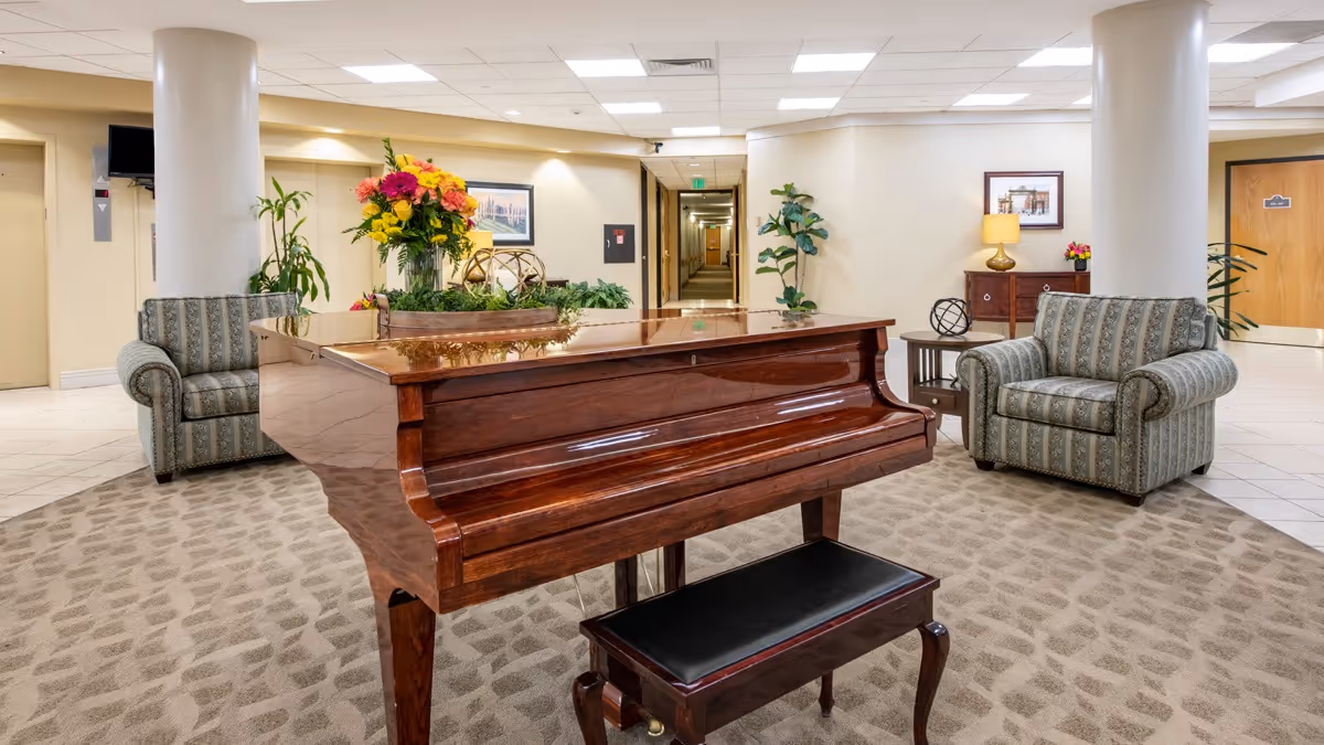 A spacious and well-lit common area in a senior living facility featuring a polished wooden grand piano with a black bench, two patterned armchairs, a side table with a decorative sculpture, a wooden cabinet with a lamp and flowers, and a large floral arrangement on the piano. The background shows a hallway, elevator, and doors, with plants and framed artwork on the walls.