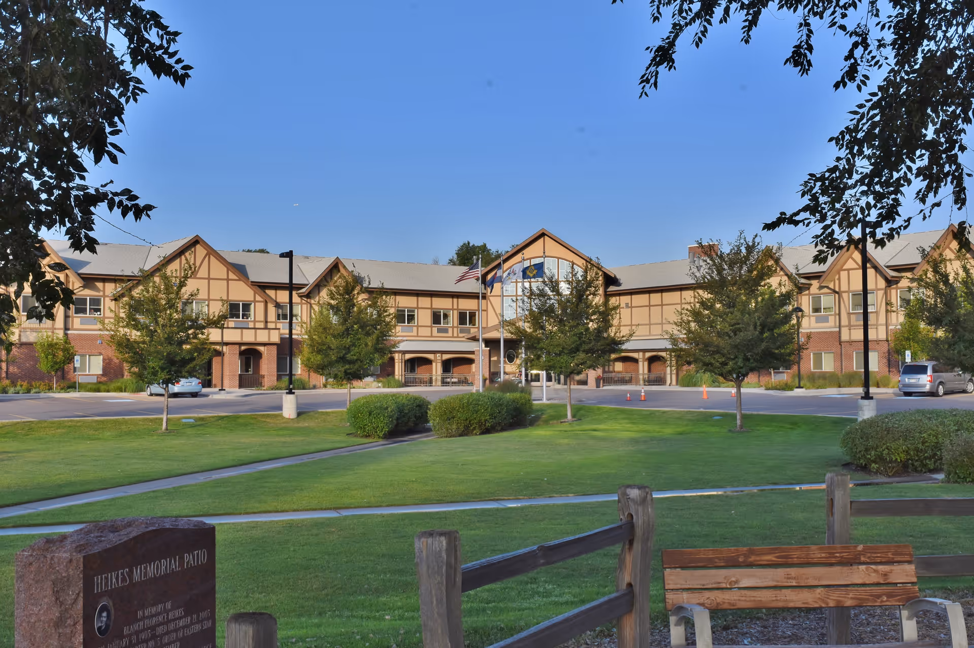 Exterior view of the Eastern Star Masonic Retirement Campus building with a well-maintained lawn, trees, and a wooden bench in the foreground under a clear blue sky.