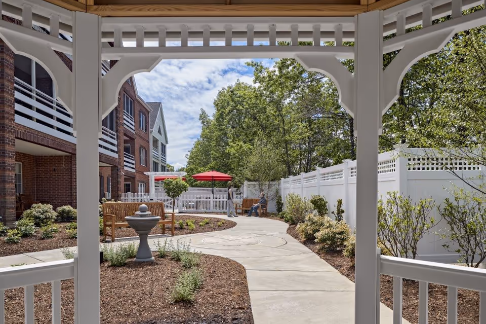 View from inside a white gazebo looking out onto a curved concrete pathway in a garden area with benches, a small fountain, and landscaping. Two people are sitting and standing near a bench under a red umbrella. The background shows a multi-story brick building and trees under a partly cloudy sky.