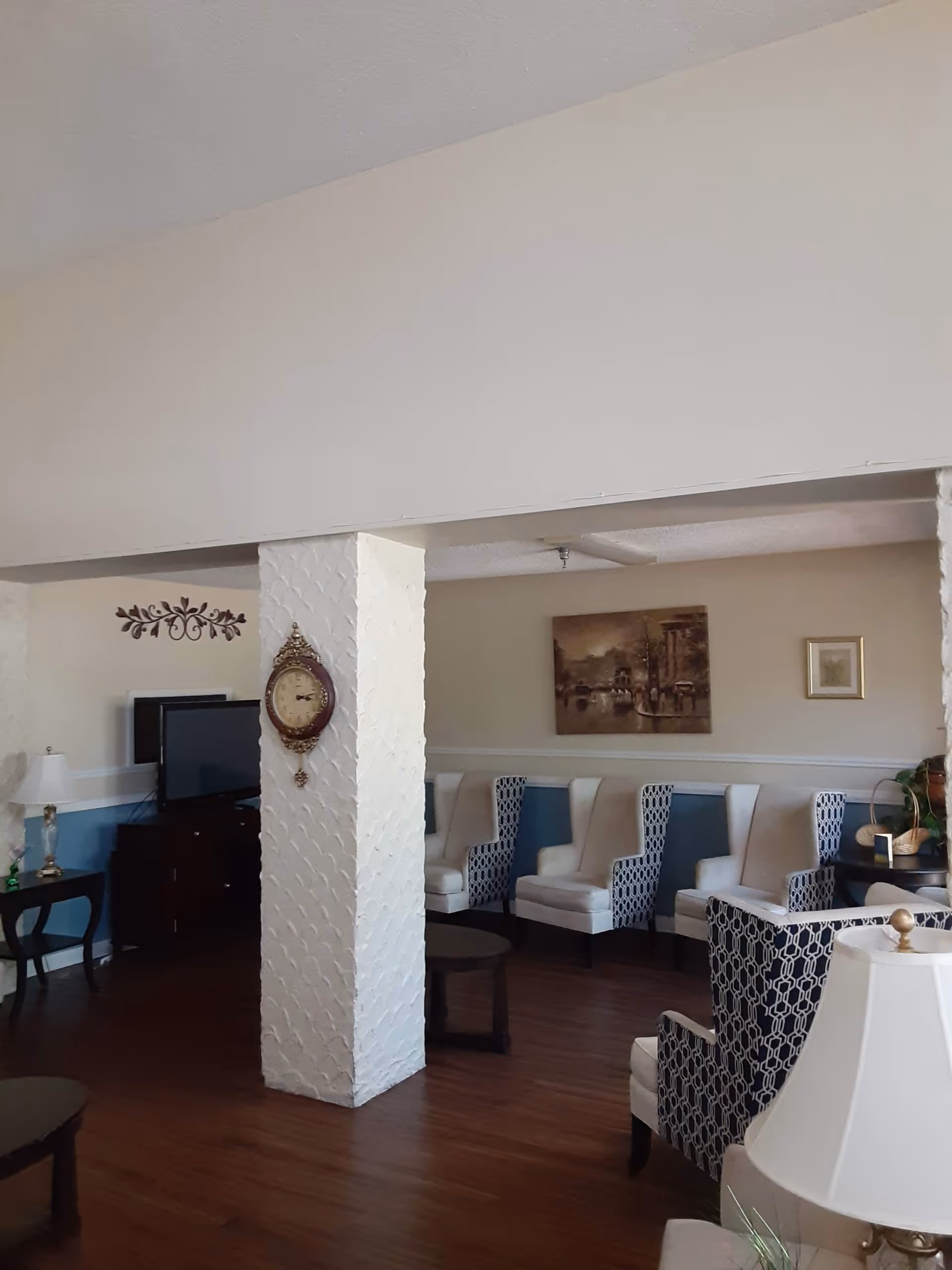 A cozy living room area with wooden flooring, several upholstered armchairs with patterned backs, a white textured pillar with a decorative clock mounted on it, a flat-screen TV on a dark wooden stand, a side table with a lamp, and framed artwork on the walls.