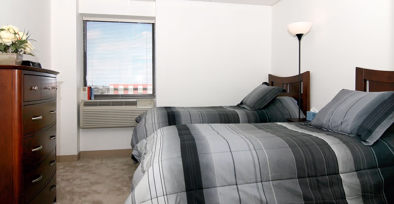 Two twin beds with gray-striped bedding in a simple bedroom with a dresser, window air conditioner, and floor lamp.