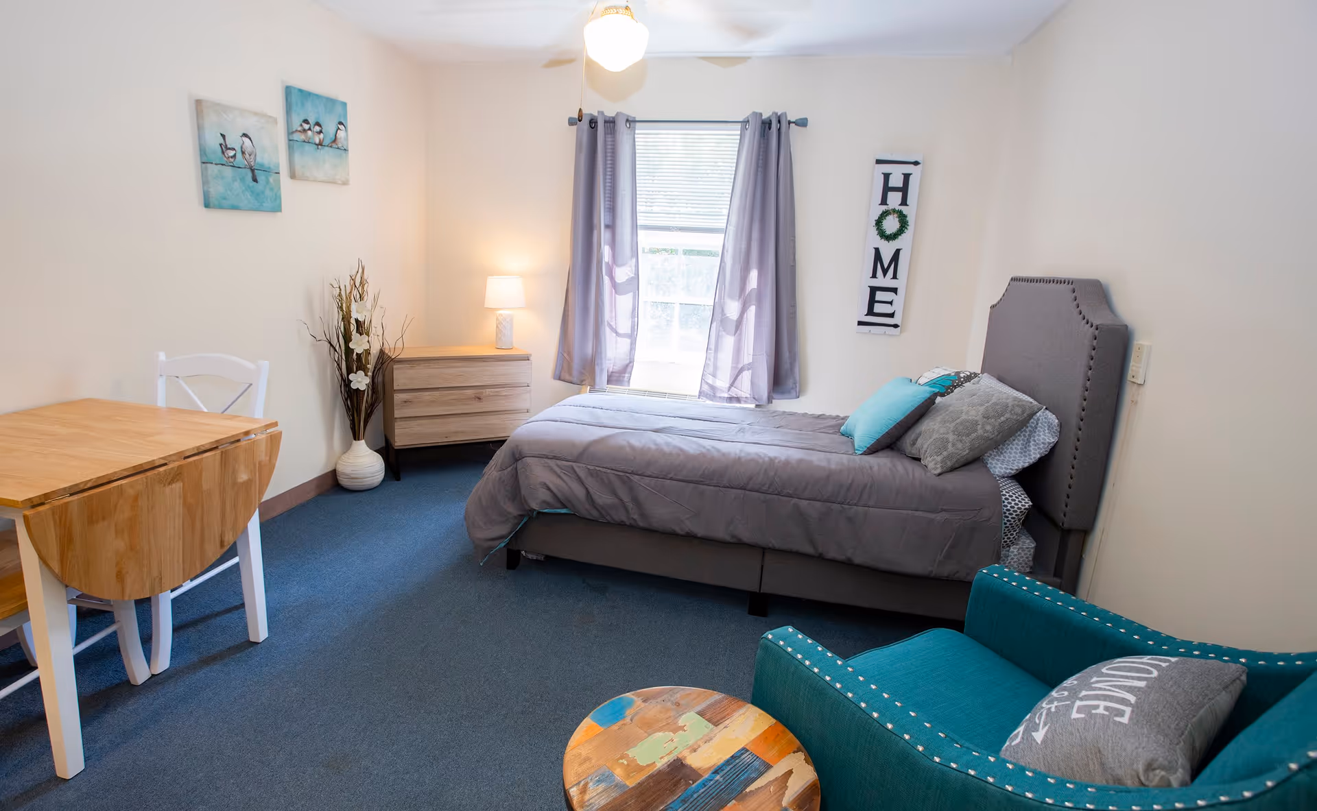 A cozy bedroom with a single bed made up with gray bedding and multiple pillows, a teal armchair with a decorative pillow, a small round wooden table, a wooden drop-leaf table with a white chair, a wooden dresser with a lamp, a window with gray curtains, and wall decorations including two bird paintings and a vertical 'HOME' sign.