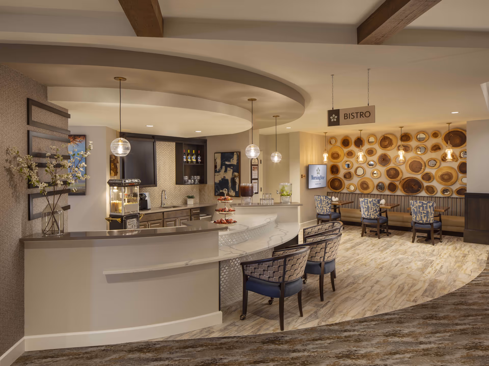 Interior view of a modern bistro area in MorningStar Assisted Living & Memory Care of Rio Rancho featuring a curved counter with three chairs, pendant lights hanging from the ceiling, a popcorn machine, and a seating area with tables and chairs against a wall decorated with circular wooden art pieces and mirrors.
