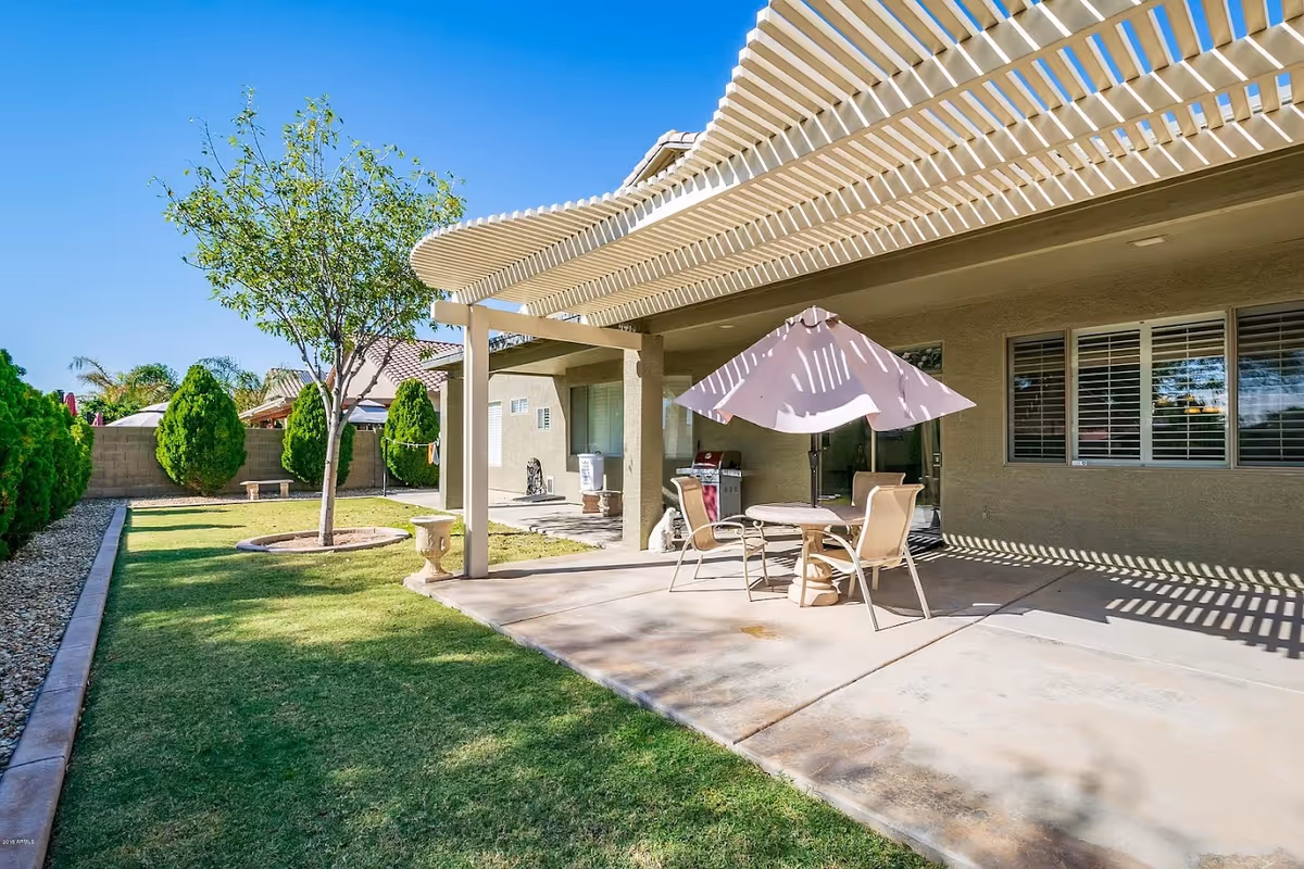 Outdoor patio area with a pergola providing partial shade over a round table with four chairs and a large umbrella. The patio is adjacent to a beige building with windows and a sliding door. The yard features a green lawn, a small tree, decorative urns, and a stone bench along a stone-lined border. The sky is clear and blue.