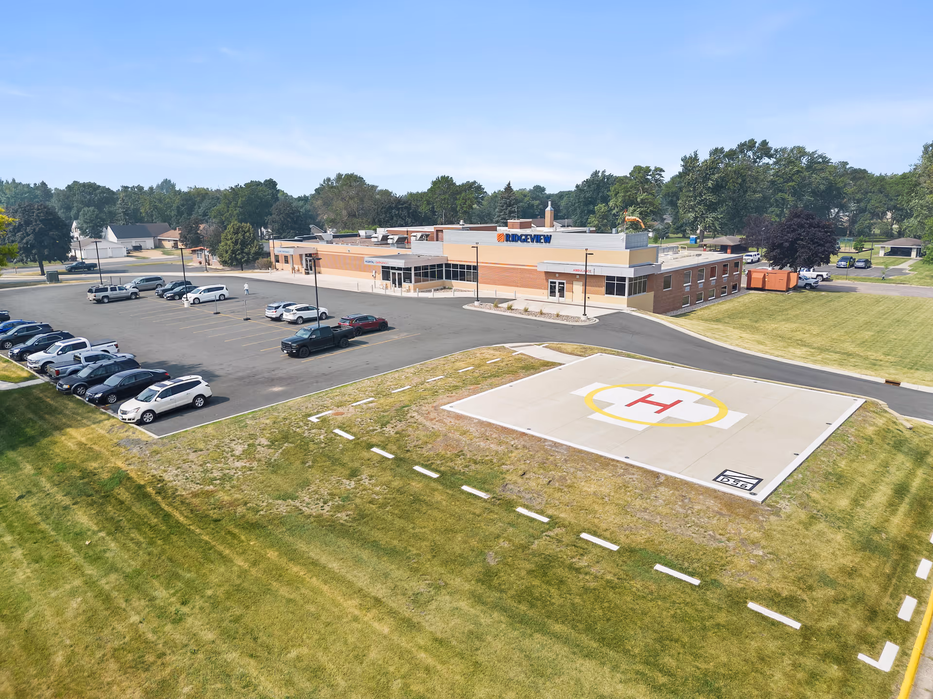 Aerial view of a single-story brick building with a parking lot filled with cars and a helipad in the foreground. The building has a sign that reads 'RIDGEVIEW' and is surrounded by green grass and trees under a clear blue sky.