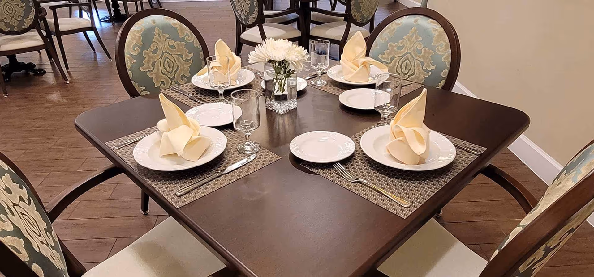 A dining table set for four with white plates, folded beige napkins, clear drinking glasses, silverware, and a small vase with white flowers in the center. The table is dark wood with woven placemats, surrounded by upholstered chairs with a blue and beige patterned fabric. Additional dining tables and chairs are visible in the background on a wooden floor.