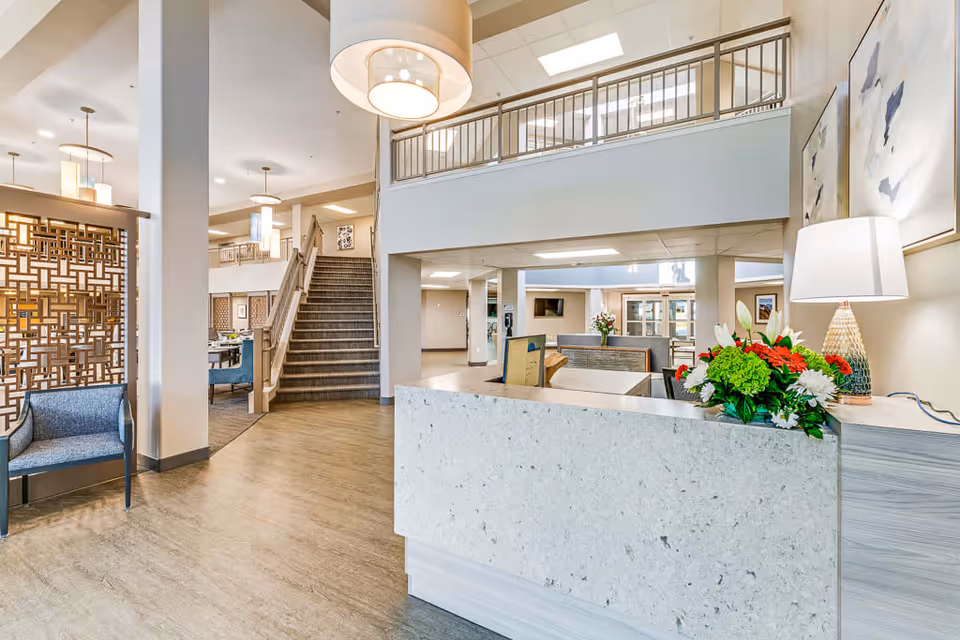 Bright and spacious assisted living facility interior with a reception desk adorned with a floral arrangement and a lamp. To the left, there is a blue armchair and decorative wooden partition. In the background, a staircase leads to an upper level with railings, and dining area seating is visible. The space is well-lit with modern hanging light fixtures and neutral-toned walls and flooring.