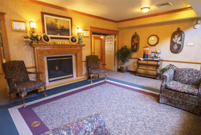 A cozy living room area with floral upholstered chairs and a sofa arranged around a fireplace with a wooden mantel. The mantel is decorated with framed pictures, plates, and a floral arrangement. The walls are adorned with framed artwork and a clock. There is a small wooden chest with a water dispenser and fruit basket on top, and a potted plant in the corner. The room has warm lighting and a patterned carpet with purple and blue borders.