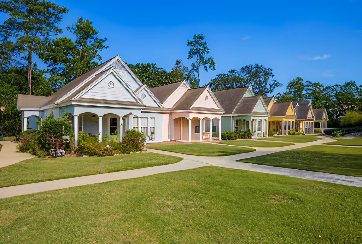 Row of single-story cottage-style residences with covered porches along walkways and manicured lawns under a clear blue sky.