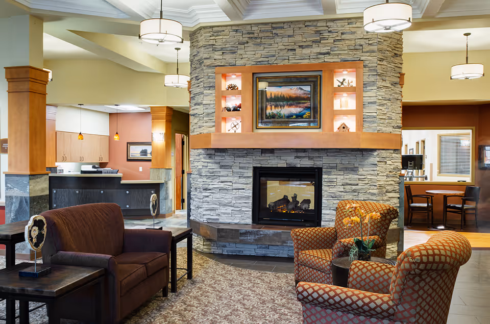 A cozy senior living facility common area with a stone fireplace in the center, decorated with a framed landscape painting and small decorative items on built-in shelves. The room features comfortable seating including a brown loveseat and two patterned armchairs around a small table with a flower arrangement. The background shows a reception desk and a small dining area with tables and chairs. The space is warmly lit with ceiling lights and pendant lamps.