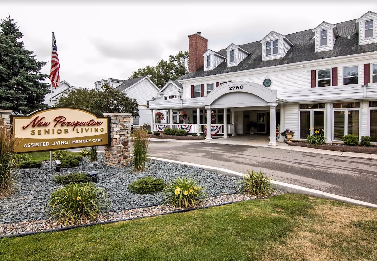 Exterior view of New Perspective Senior Living facility in Roseville, showing a large white building with multiple windows and dormers, a covered entrance with columns, and a landscaped area with a sign that reads 'New Perspective Senior Living Assisted Living | Memory Care'. An American flag is also visible near the sign.