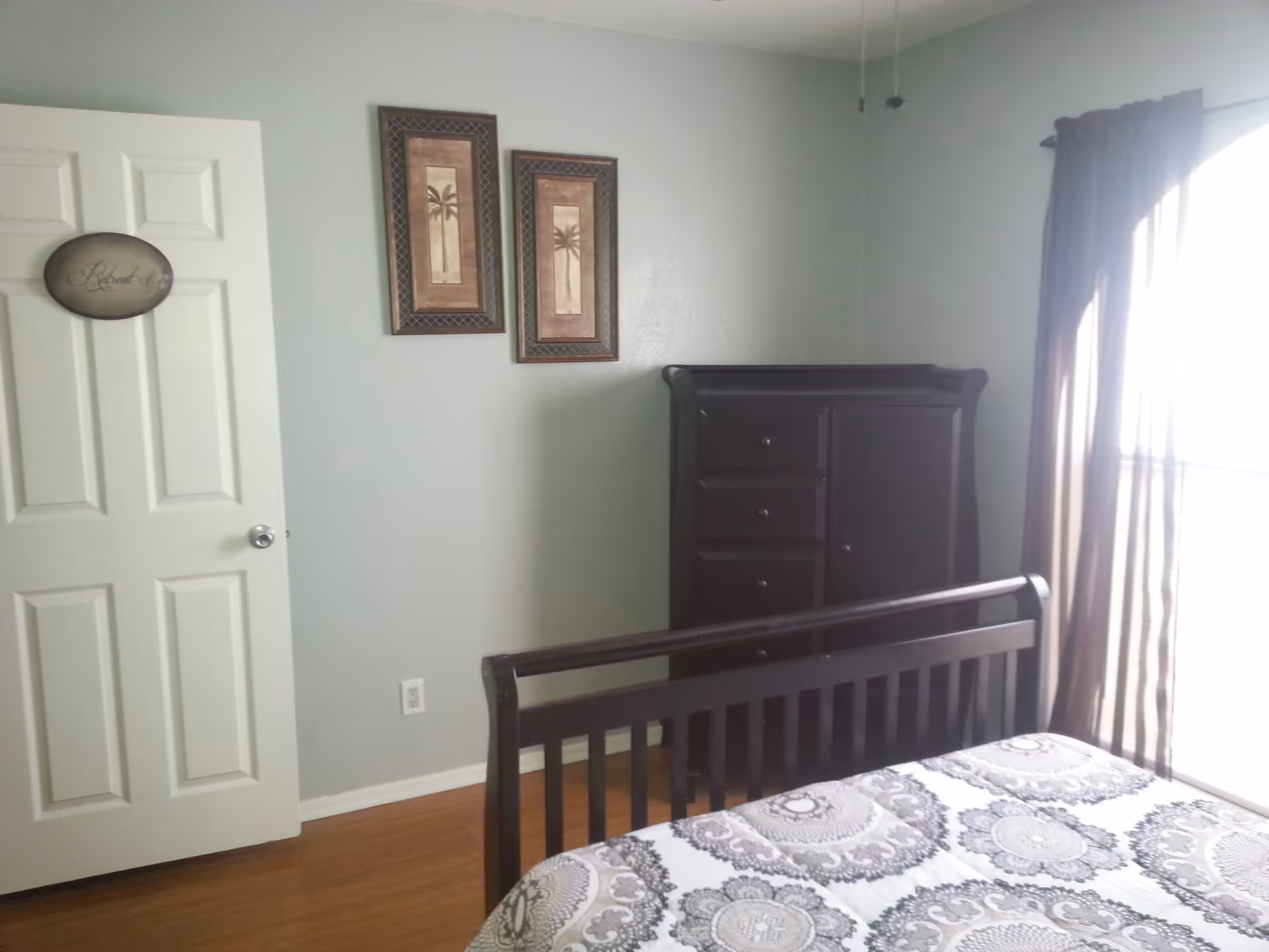 A bedroom with a wooden bed frame and patterned bedspread, a dark wooden dresser, two framed palm tree pictures on a light green wall, a white door with a sign that says 'Retreat', and a window with sheer brown curtains letting in sunlight.
