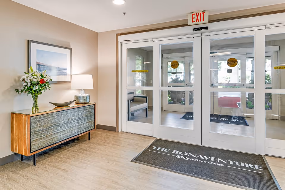 Interior view of an assisted living facility entrance area featuring automatic glass doors with an exit sign above. To the left, there is a wooden console table with a vase of flowers, a decorative bowl, and a lamp. A framed picture hangs on the wall above the table. A floor mat in front of the doors reads 'THE BONAVENTURE SKY ACTIVE LIVING.'
