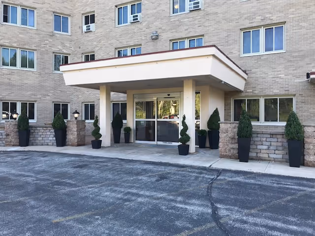 Entrance of a multi-story brick building with a covered drop-off area supported by two columns. Several potted plants and small trees line the entrance area. Multiple windows are visible on the building facade.