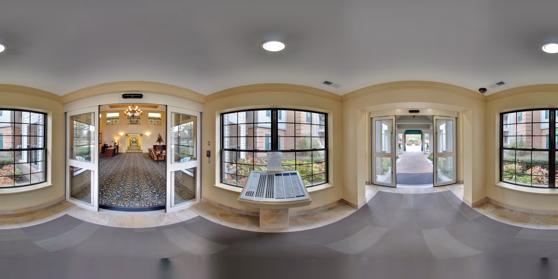 Panoramic view of the entrance area inside Oakmont Manor Senior Community showing double glass doors leading to a carpeted hallway with chandeliers and furniture. Large windows on either side reveal outdoor landscaping with plants and brick buildings.