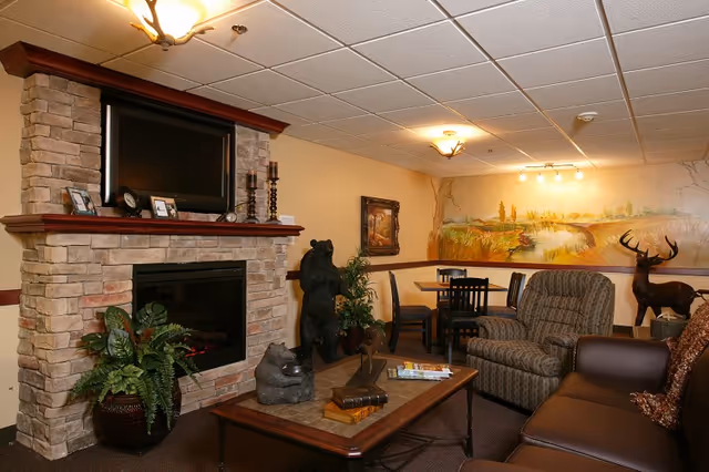 Cozy common room with a stone fireplace and mounted TV, sofa and armchairs around a coffee table, a small dining table in the back, and a nature-themed mural on the wall.