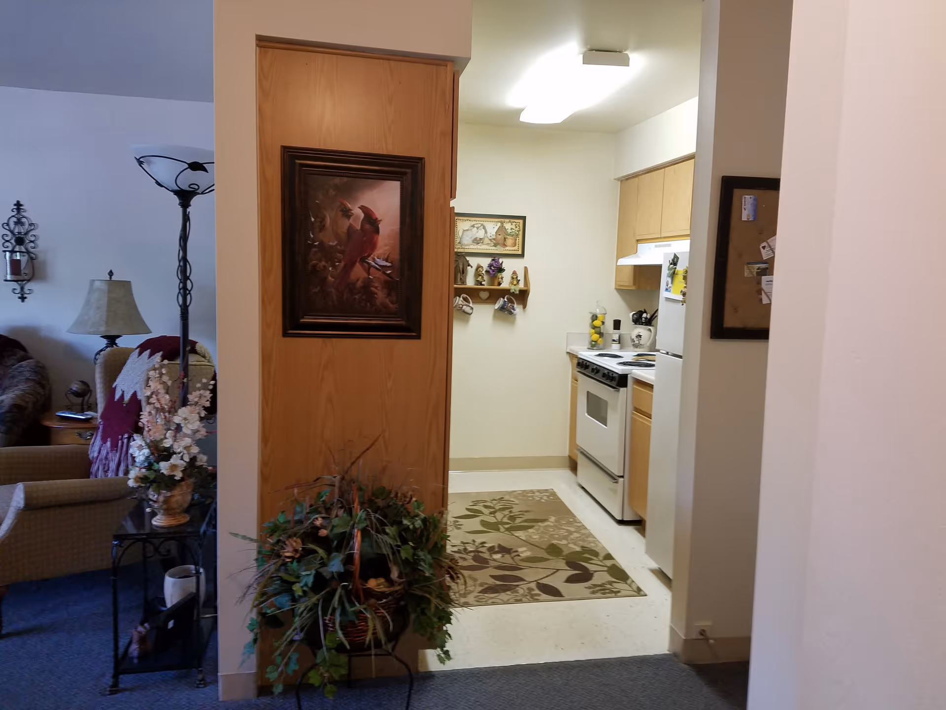 View of a small kitchen area with wooden cabinets, a white stove, and a refrigerator. A decorative rug with leaf patterns is on the floor. To the left, part of a living room is visible with a beige armchair, a floor lamp, a table lamp, and floral decorations. A framed painting of a cardinal bird is mounted on a wooden panel between the kitchen and living room areas.