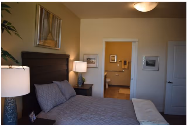 A bedroom with a neatly made bed featuring gray bedding and pillows. There are two bedside tables with lamps on either side of the bed. A framed picture hangs above the headboard. The room has beige walls and a door that leads to a bathroom, where a toilet and towel rack are visible.