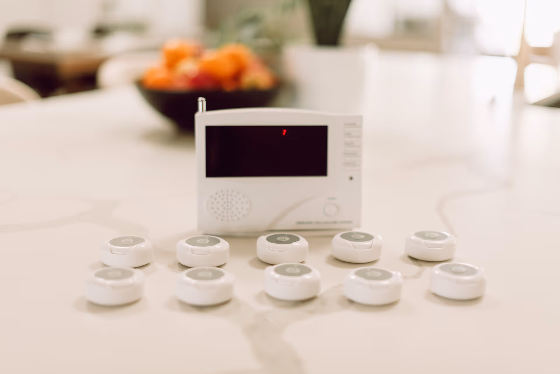 A white emergency call system device with a screen and speaker, placed on a white marble countertop. In front of the device are ten small white circular call buttons arranged in two rows. In the background, there is a blurred bowl of fruit and some greenery.