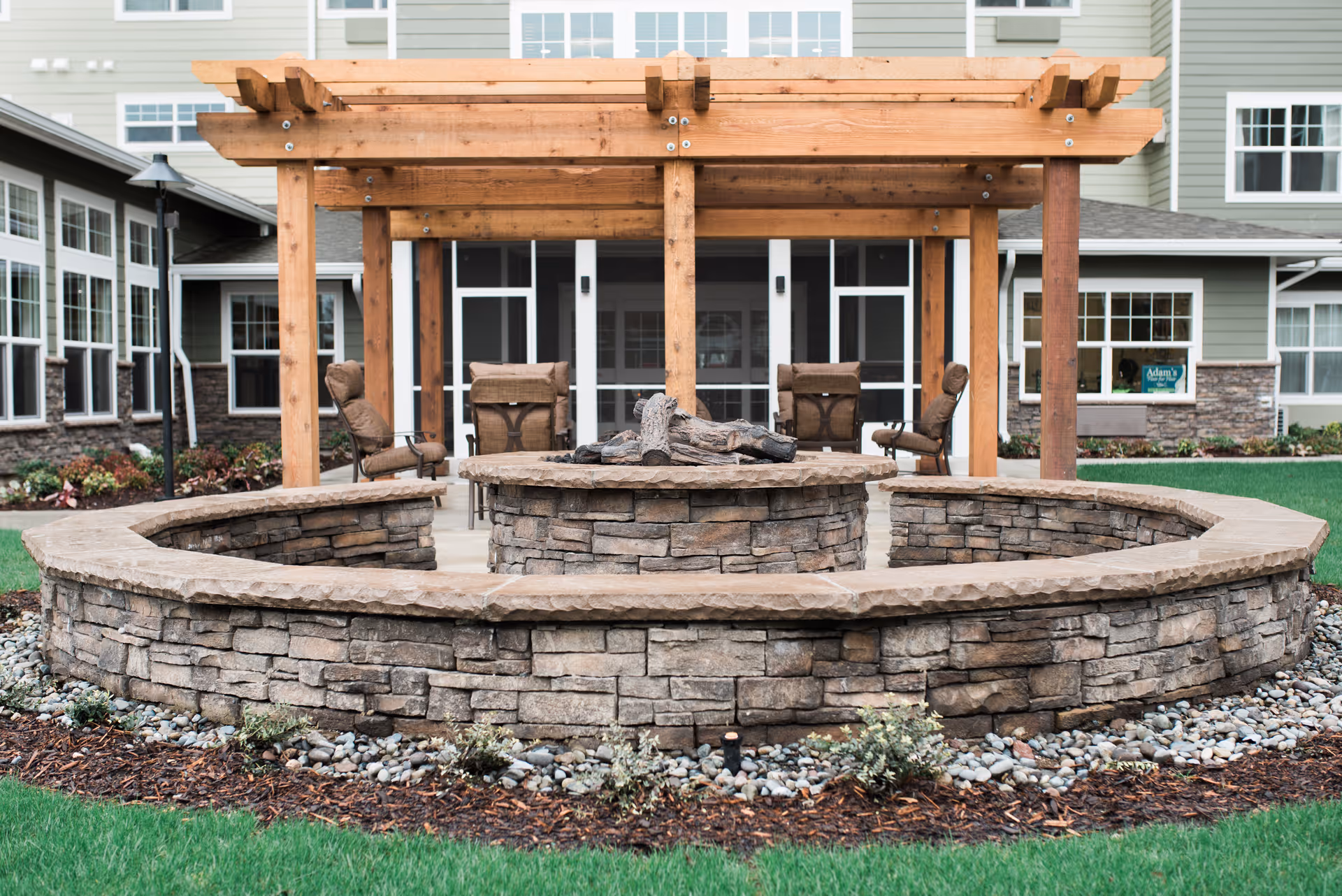 Outdoor seating area with a circular stone fire pit surrounded by a stone wall and cushioned chairs under a wooden pergola, with a building featuring large windows in the background.