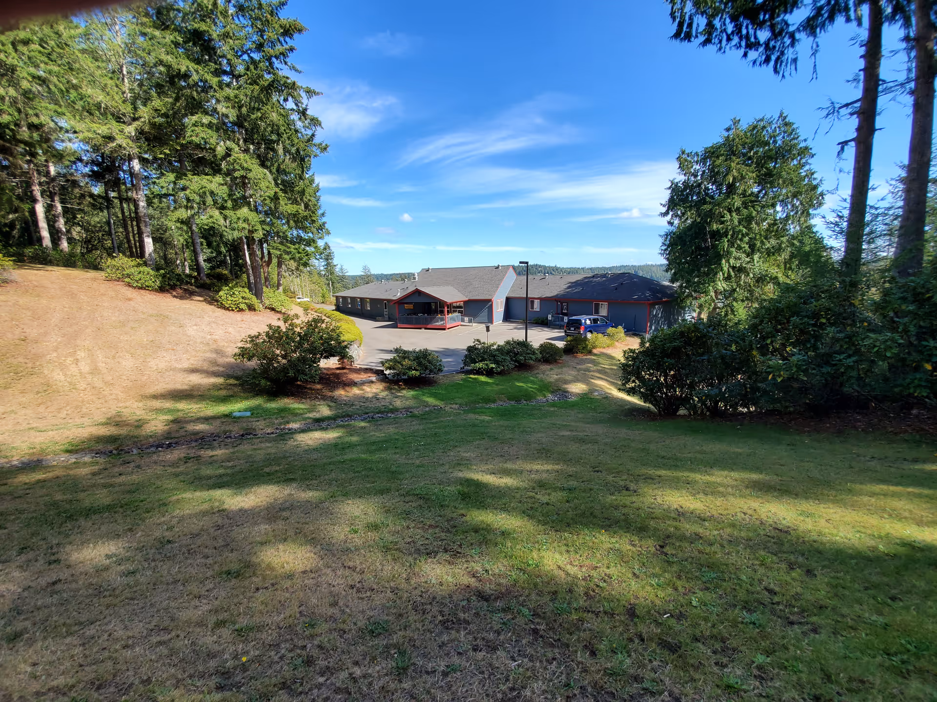 A single-story building with a gray roof and blue-gray walls surrounded by trees and bushes under a clear blue sky. The building is situated at the end of a paved area with a few parked cars, and there is a grassy area with some shadows in the foreground.