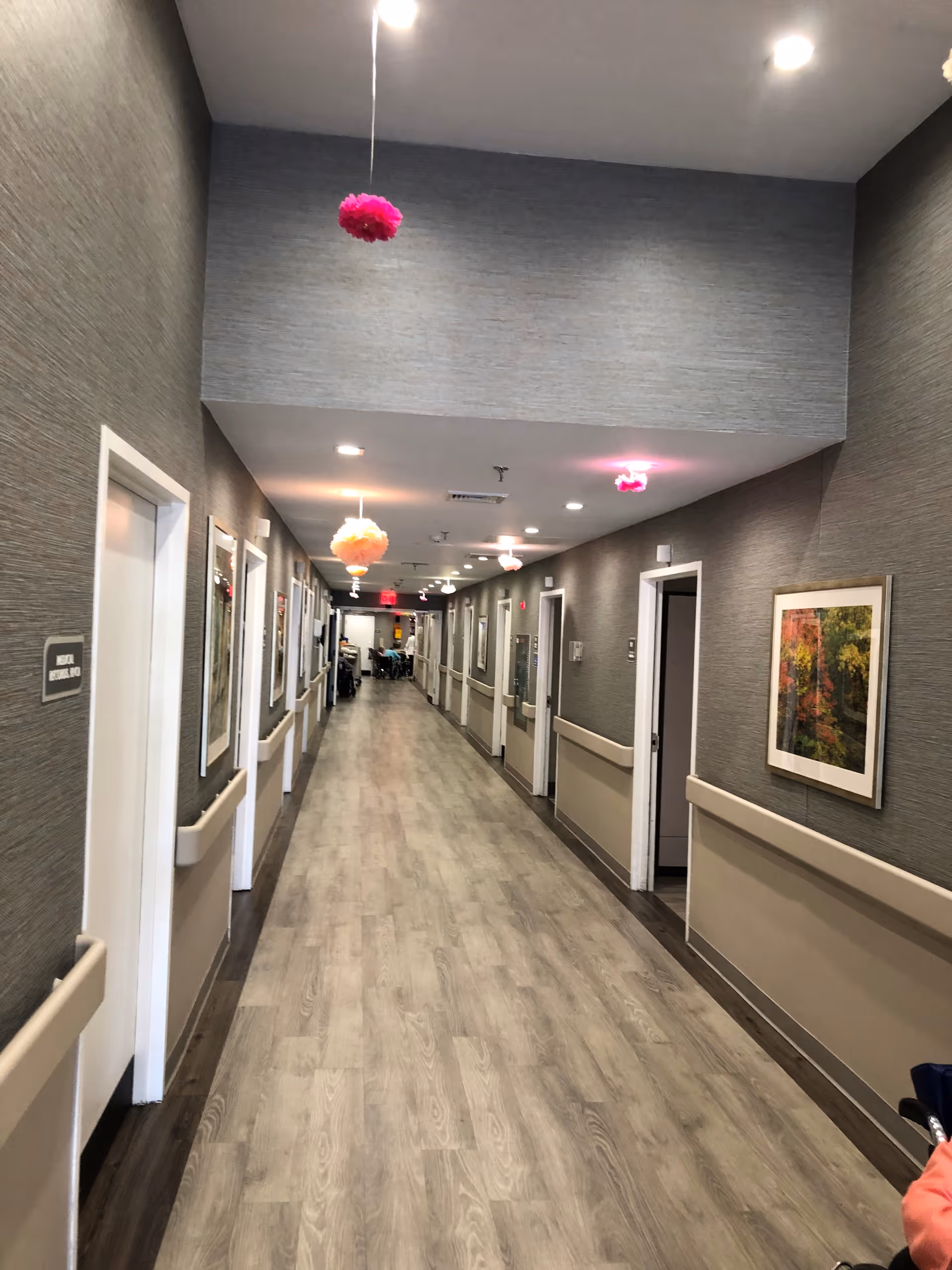 A long, well-lit hallway in a senior living facility with wood-patterned flooring and gray textured walls. Several doors line both sides of the hallway, with handrails along the walls. Colorful hanging decorations are suspended from the ceiling, and framed artwork is displayed on the walls. At the far end of the hallway, some people are visible sitting at a table.