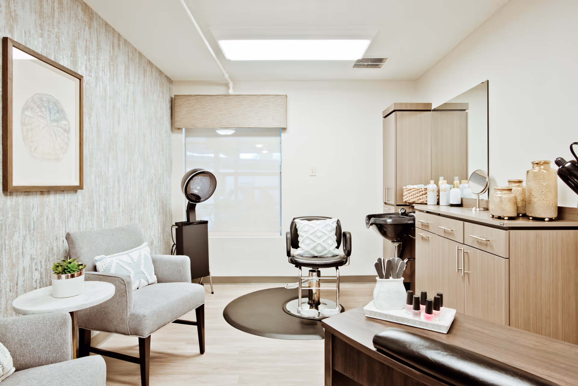 A bright and clean salon area in a senior living facility featuring a styling chair with a cushion, a hair dryer, a small sink, wooden cabinets with various hair care products, and two gray armchairs with pillows next to a small round table with a potted plant. The room has light-colored walls and flooring, with a framed artwork on one wall.