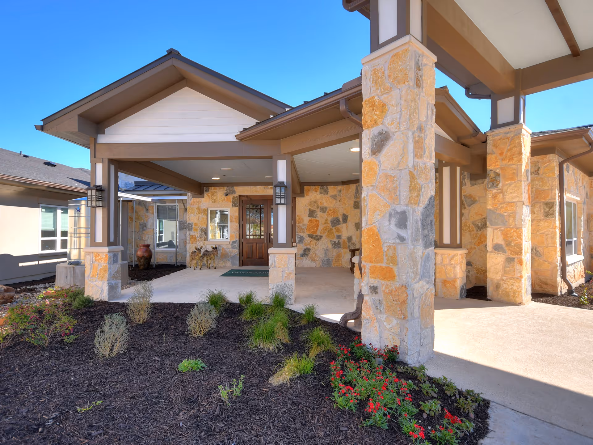 Exterior view of the entrance to Alexis Pointe of Wimberley Senior Living facility featuring stone pillars, a covered porch, landscaped garden beds with small plants and flowers, and a wooden front door under a clear blue sky.