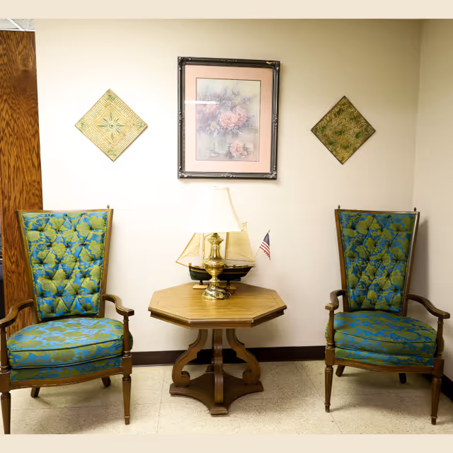Two vintage-style armchairs with blue and green floral upholstery are positioned on either side of a wooden octagonal side table. On the table is a brass lamp with a white shade and a small model sailboat with an American flag. The wall behind features a framed floral painting in the center and two decorative square plaques on either side.