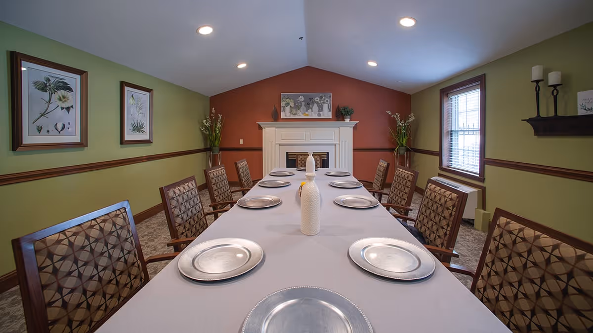 A dining room with a long table covered with a white tablecloth, set with eight silver plates and a white decorative vase in the center. The room has green walls with a brown trim, two framed botanical prints on the left wall, a window with blinds on the right, and a white fireplace with a floral painting above it at the far end. There are patterned chairs around the table and two tall plants flanking the fireplace.