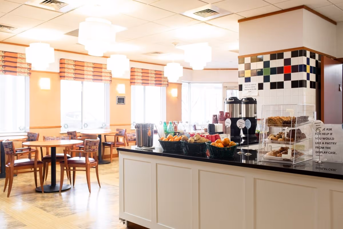 Bright dining area with round tables and chairs and a self-serve counter offering coffee, fruit, and pastries.