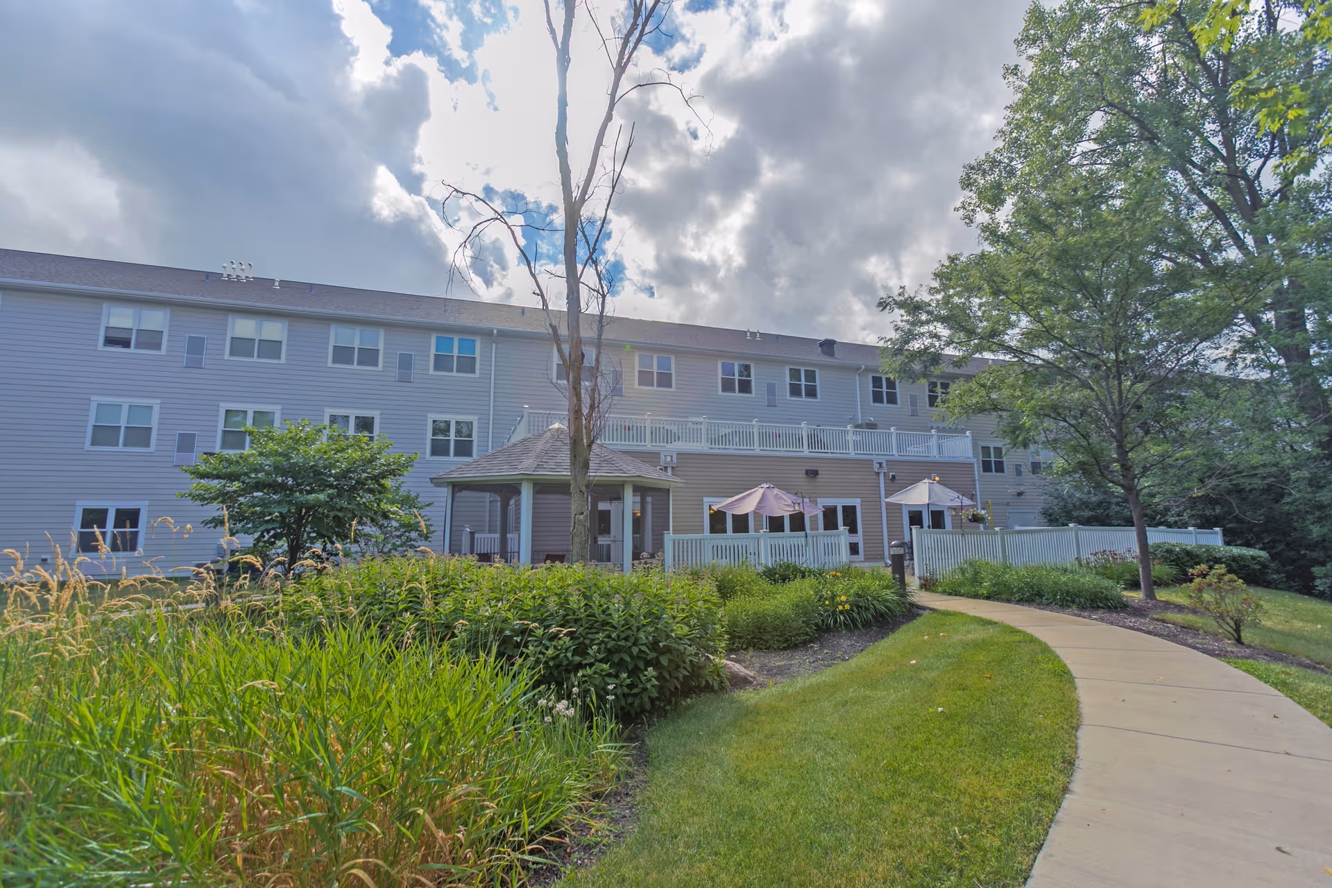 Exterior view of a multi-story senior living facility building with light gray siding, surrounded by green trees and bushes. A paved walkway curves through a well-maintained lawn leading to a small gazebo and a fenced patio area with umbrellas. The sky is partly cloudy with sunlight breaking through.