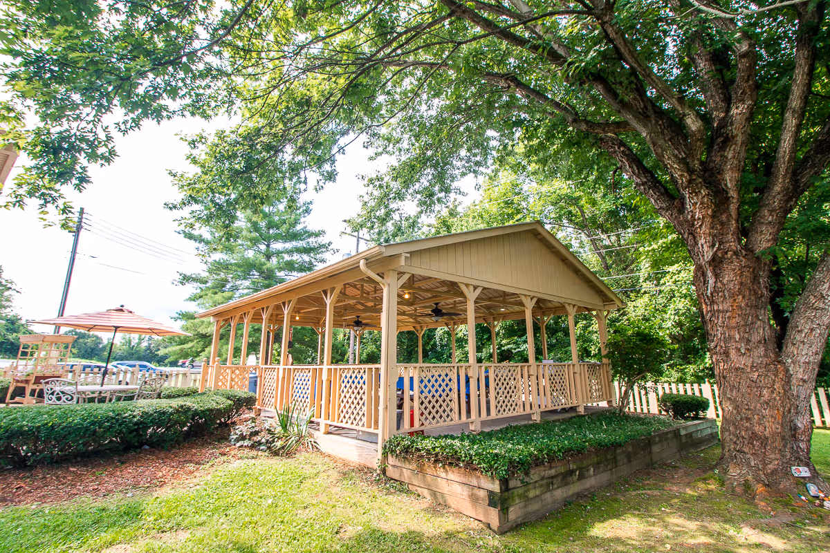 A wooden gazebo with a lattice railing situated in a green outdoor area surrounded by trees and bushes. There is a large tree to the right and an umbrella-covered seating area visible in the background on the left.