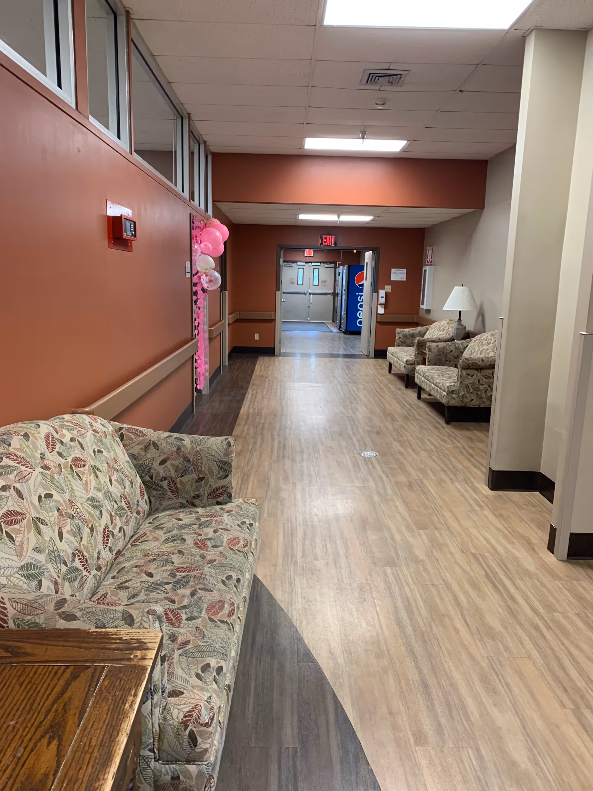 A hallway in Worthington Healthcare Center with patterned sofas and armchairs along the walls. The floor has a wood-like finish with a darker curved section near the left side. The walls are painted in a reddish-brown and beige color scheme. There are pink and white balloons attached to a door on the left side. At the end of the hallway, double doors are open, revealing a Pepsi vending machine in the next room.
