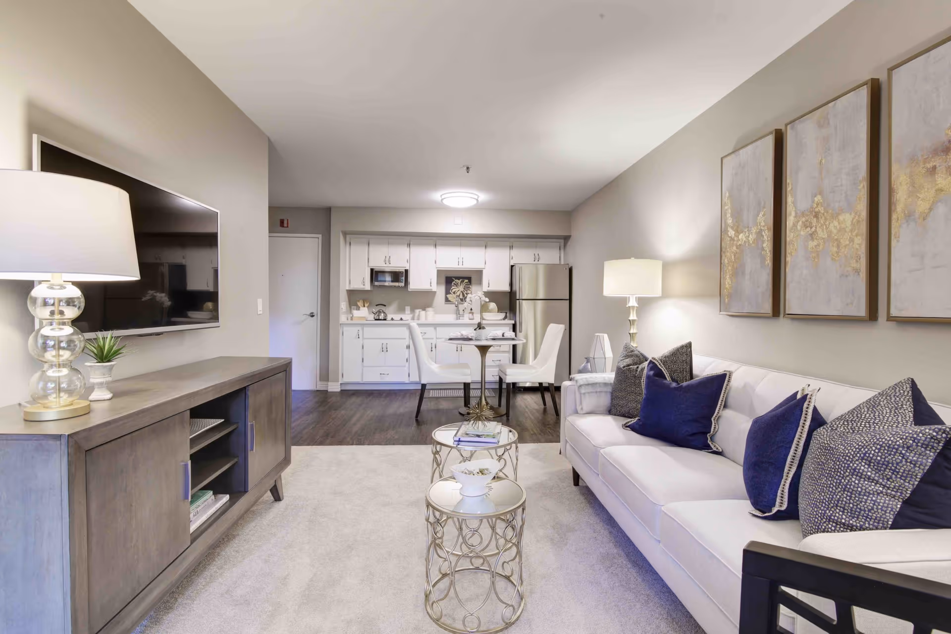 A modern senior living apartment interior featuring a white sofa with blue and patterned pillows, two round glass-top side tables, a wooden TV console with a lamp and small plant, a wall-mounted flat-screen TV, and a small dining area with a round table and two white chairs. The kitchen area in the background has white cabinets, a microwave, and a stainless steel refrigerator. The walls are painted light gray and decorated with three abstract paintings with gold accents.