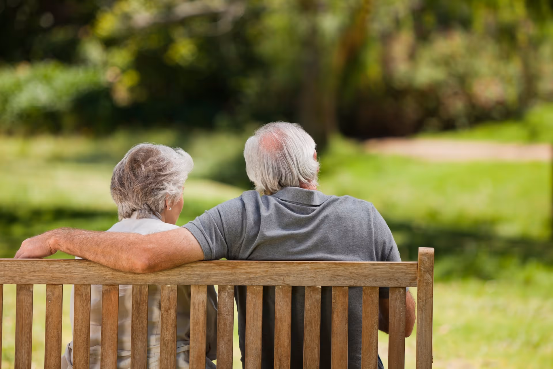 An elderly couple sitting closely together on a wooden bench in a green outdoor park area, viewed from behind.