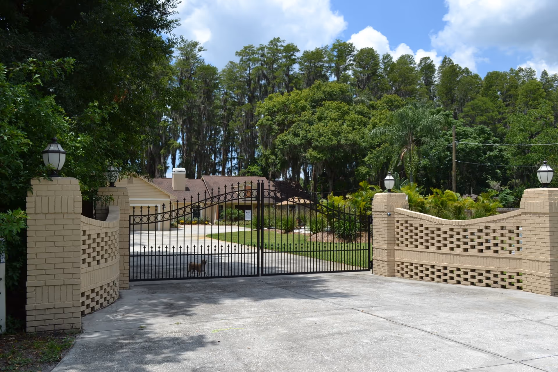 A gated entrance to Cloverdale Assisted Living featuring a black wrought iron gate flanked by beige brick pillars with lantern-style lights on top. Behind the gate, there is a driveway leading to a single-story building surrounded by lush green trees and landscaping under a partly cloudy sky.