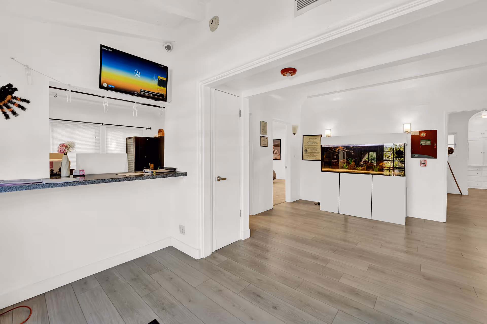 Interior view of a senior living facility reception area with a counter on the left, a wall-mounted TV above it, and a large fish tank built into a white cabinet on the right. The room has light wood flooring, white walls, and several doorways leading to other rooms.