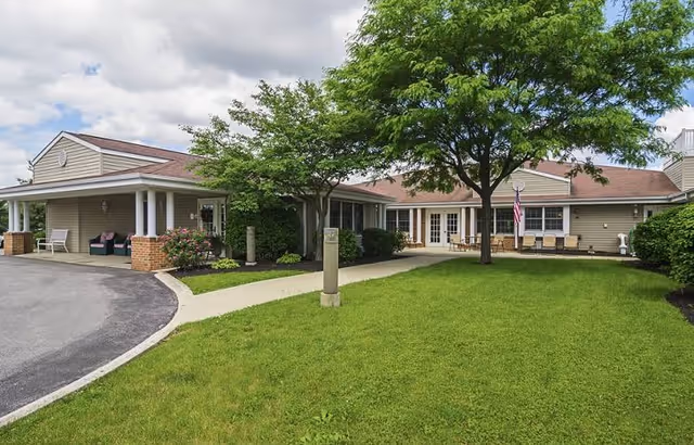 Exterior view of a single-story senior living facility building with beige siding and a red roof. There is a paved driveway on the left, a green lawn with a large tree in the center, and a concrete walkway leading to the entrance. Outdoor seating is visible near the entrance, and an American flag is displayed on the right side of the building.