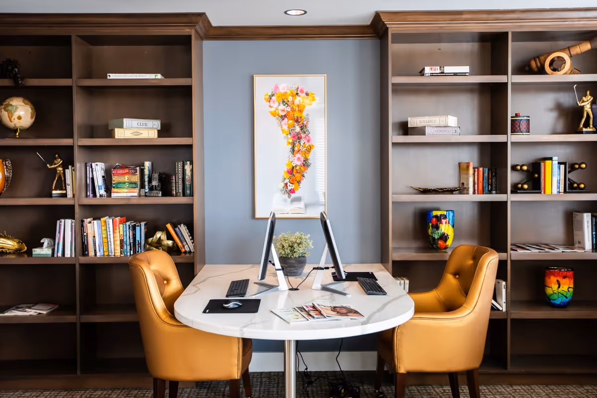 A cozy interior room with a white marble table featuring two computer monitors, keyboards, and a mouse. Two tan leather chairs are positioned on either side of the table. Behind the table, there are large wooden bookshelves filled with books, decorative items, and board games. A framed artwork of colorful flowers is centered on the blue-gray wall between the bookshelves.